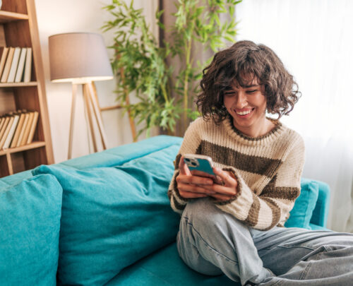 A beautiful young woman is sitting cross-legged on a couch, completely relaxed while holding a phone in both hands. Her expression shifts between focus and a soft smile, immersed in whatever she's reading or watching.